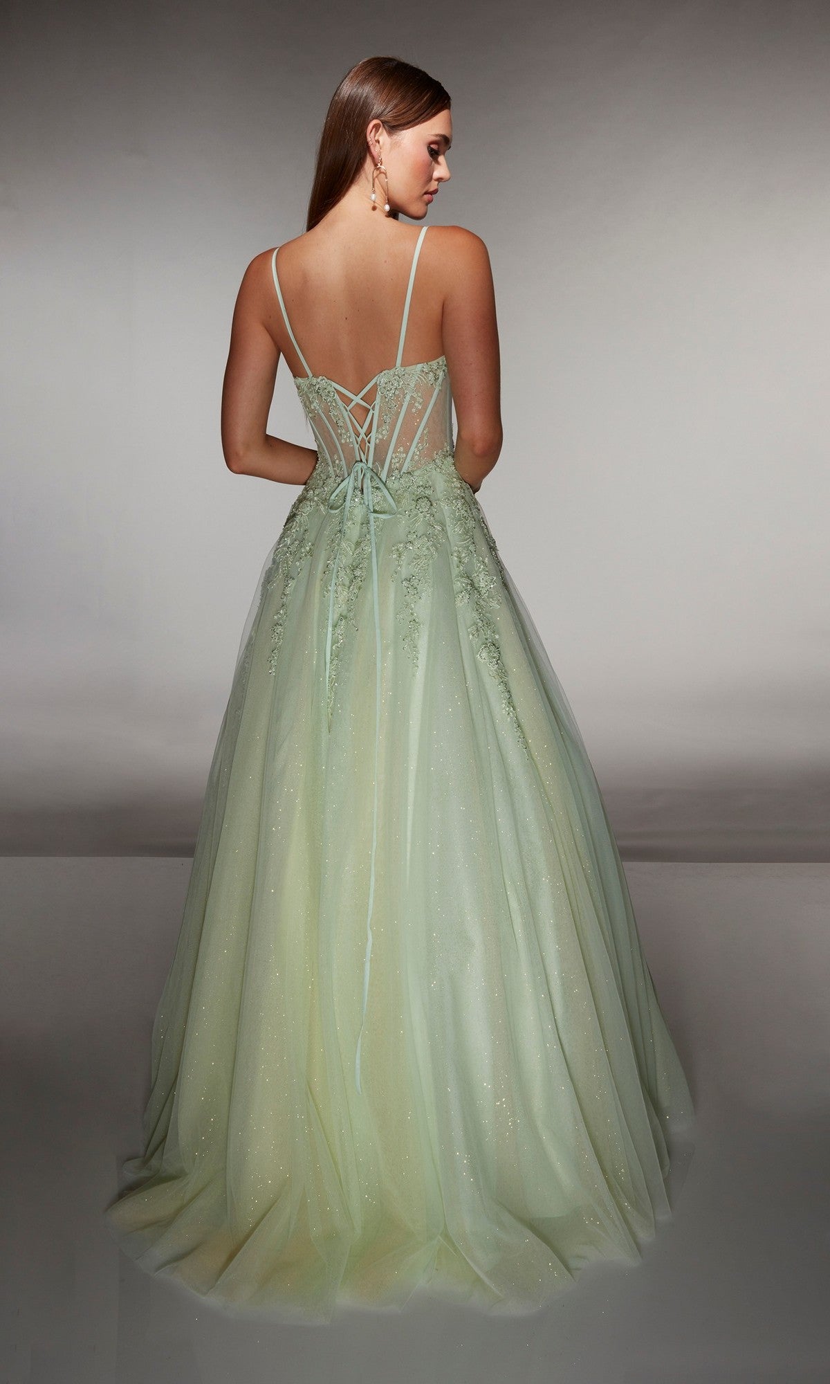 A woman poses in the Alyce 62061 long prom dress—a light green, sparkly sleeveless gown with a lace corset bodice, thin straps, and a lace-up semi-sheer back—against a plain background.